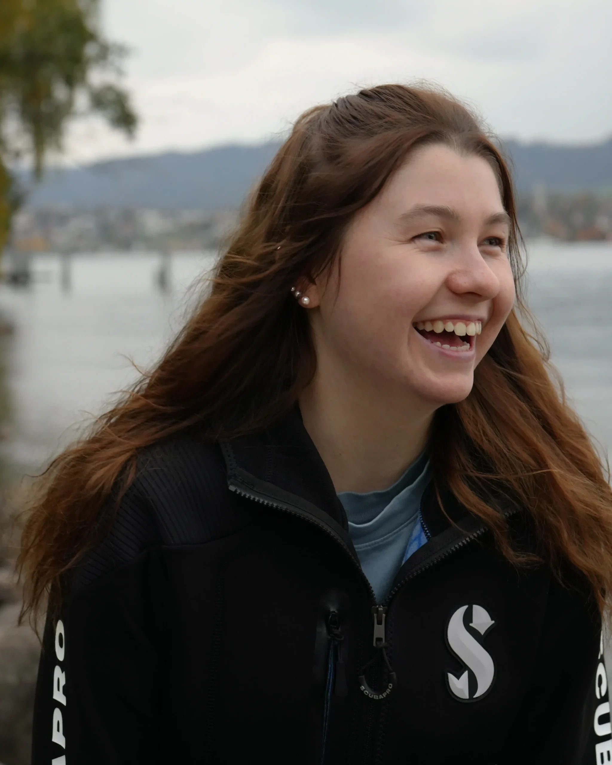 Eine junge Frau mit braunen, offenen Haaren lacht herzlich im Freien. Sie trägt eine schwarze Scubapro-Jacke und steht an einem Seeufer, im Hintergrund sind Wasser, Bäume und sanfte Hügel zu sehen. Die Stimmung ist fröhlich und natürlich.