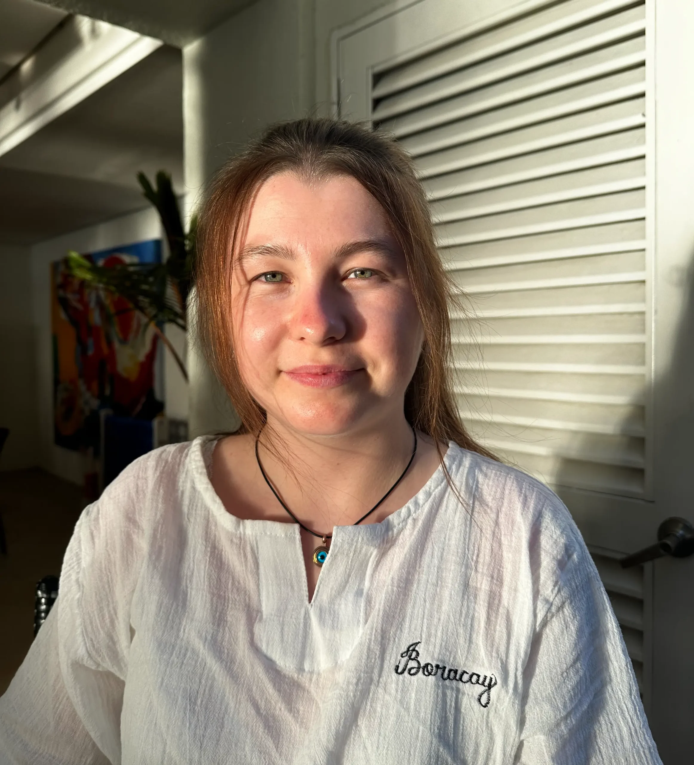 Eine junge Frau sitzt in warmem Sonnenlicht vor einer weissen Wand mit Lamellentür. Sie trägt ein weisses Oberteil mit der Aufschrift „Boracay“ und eine Halskette mit einem blauen Auge-Anhänger. Ihr Gesicht ist entspannt und leicht lächelnd, das Licht betont ihre natürliche Ausstrahlung.
