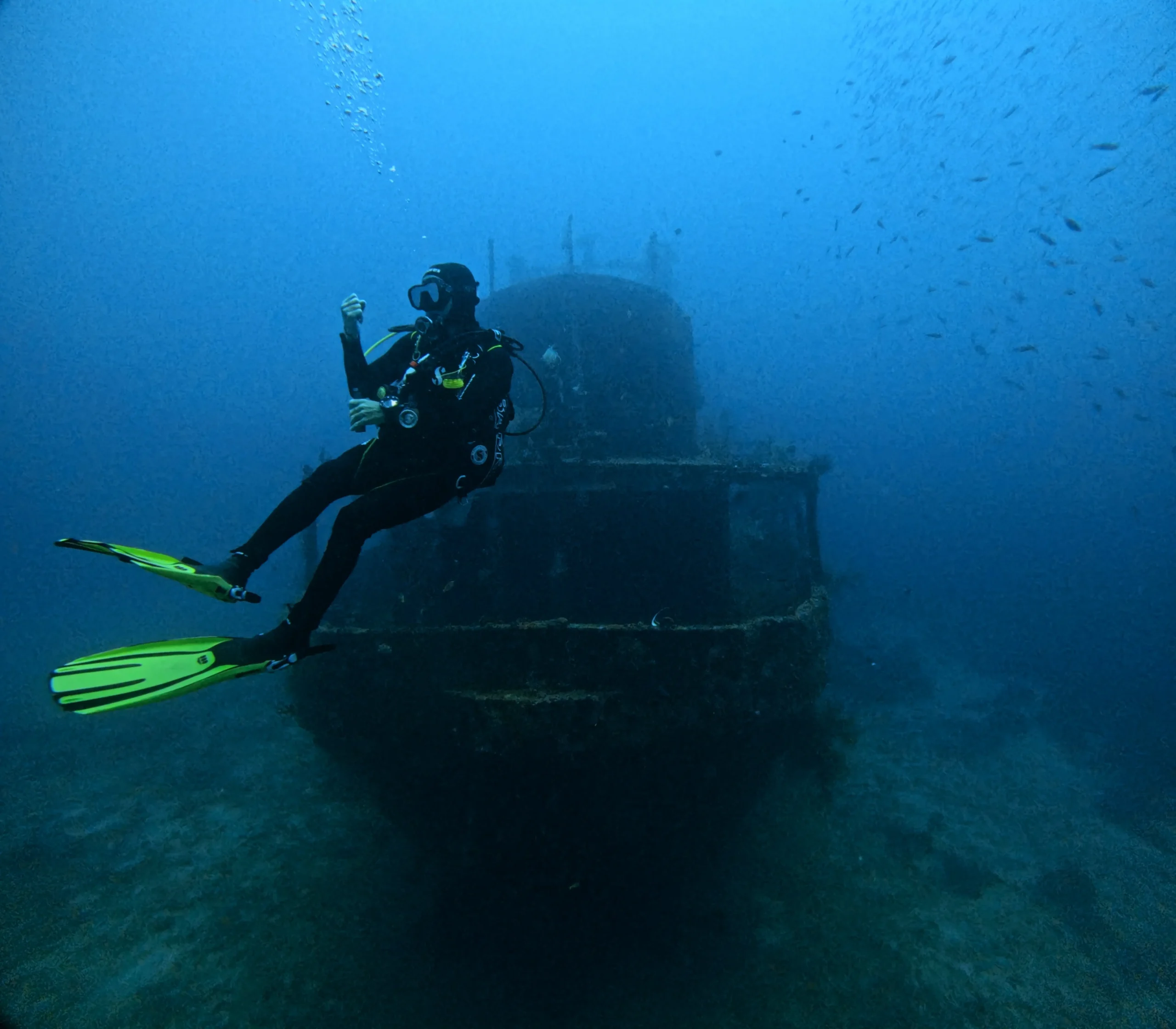 Ein Taucher mit gelben Flossen schwebt vor einem grossen Schiffswrack, das in der Tiefe ruht. Rundherum zahlreiche kleine Fische im bläulichen Wasser – typische Wracktauchsituation.